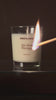A wooden match lighting the wick of the Refaire Hot Body Candle in a clear glass vessel against a warm terracotta background.